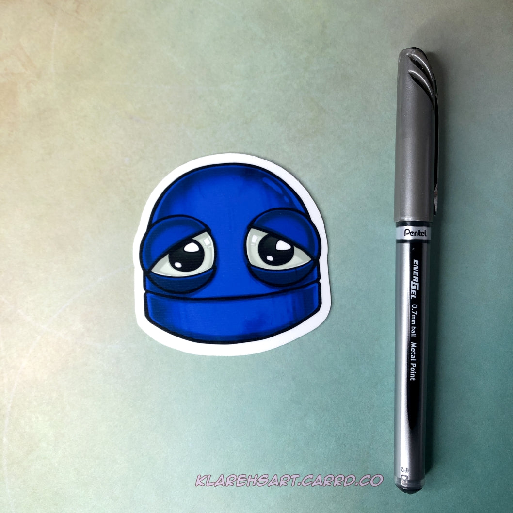 A vinyl sticker of a blue robot with a sad expression. It rests on a light gold and green background. An ink pen next to it shows size comparison.