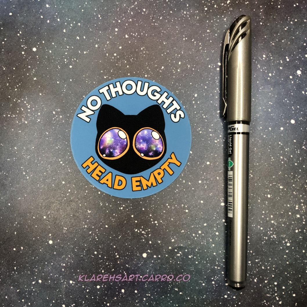 Light blue circular sticker with a black cat with galaxy eyes and text 'No Thoughts Head Empty' on a starry background. Pen next to it to show size comparison.