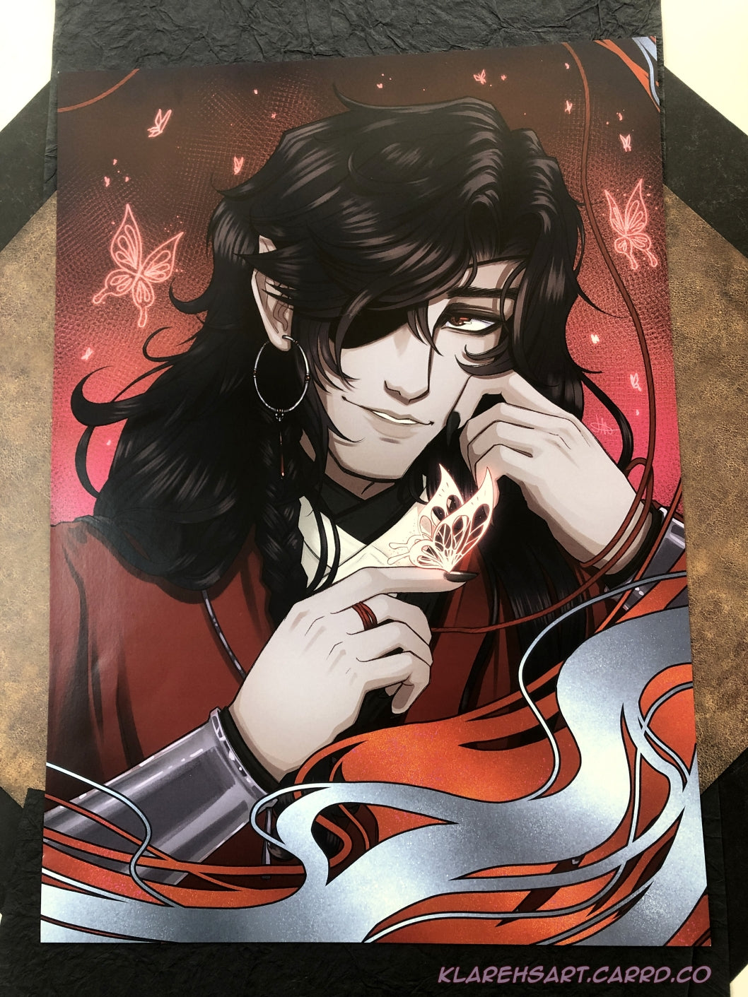 Illustration of a man with long black hair and eyepatch holding a glowing silver butterfly on his fingertip. He is wearing red, white, and black, with silver jewelry. A red background with more glowing butterflies surround him. This is the Large Size.
