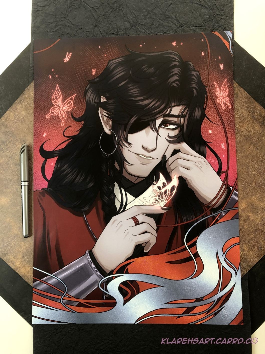 Illustration of a man with long black hair and eyepatch holding a glowing silver butterfly on his fingertip. He is wearing red, white, and black, with silver jewelry. A red background with more glowing butterflies surround him. The ink pen on the left shows size comparison. This is the Large Size.