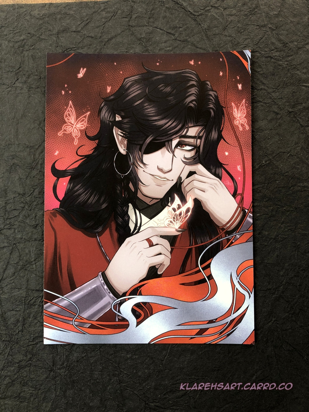 Illustration of a man with long black hair and eyepatch holding a glowing silver butterfly on his fingertip. He is wearing red, white, and black, with silver jewelry. A red background with more glowing butterflies surround him. This is the Medium Size.