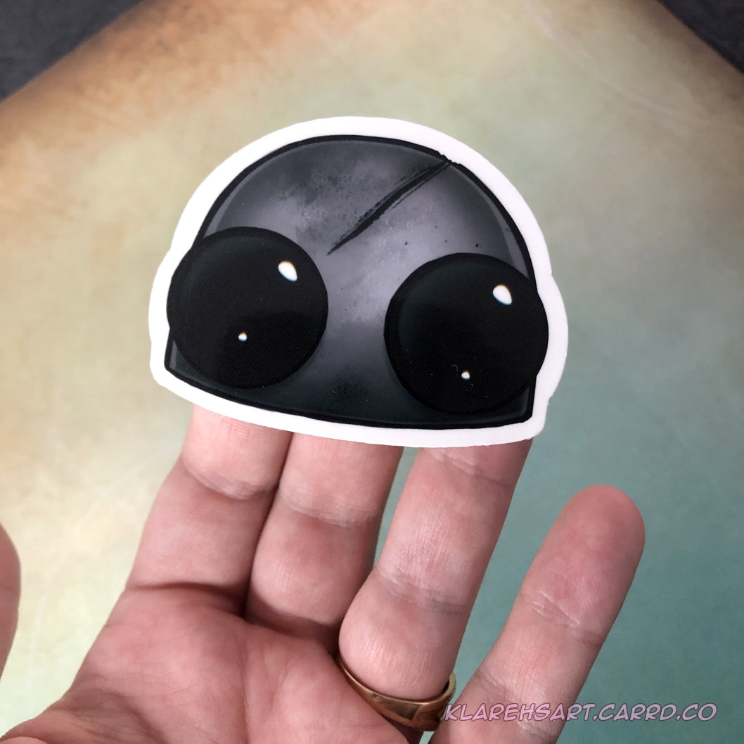 Hand holding up vinyl sticker of a dead robot head. It's hull is gray and scratched, it's eyes black with no battery.