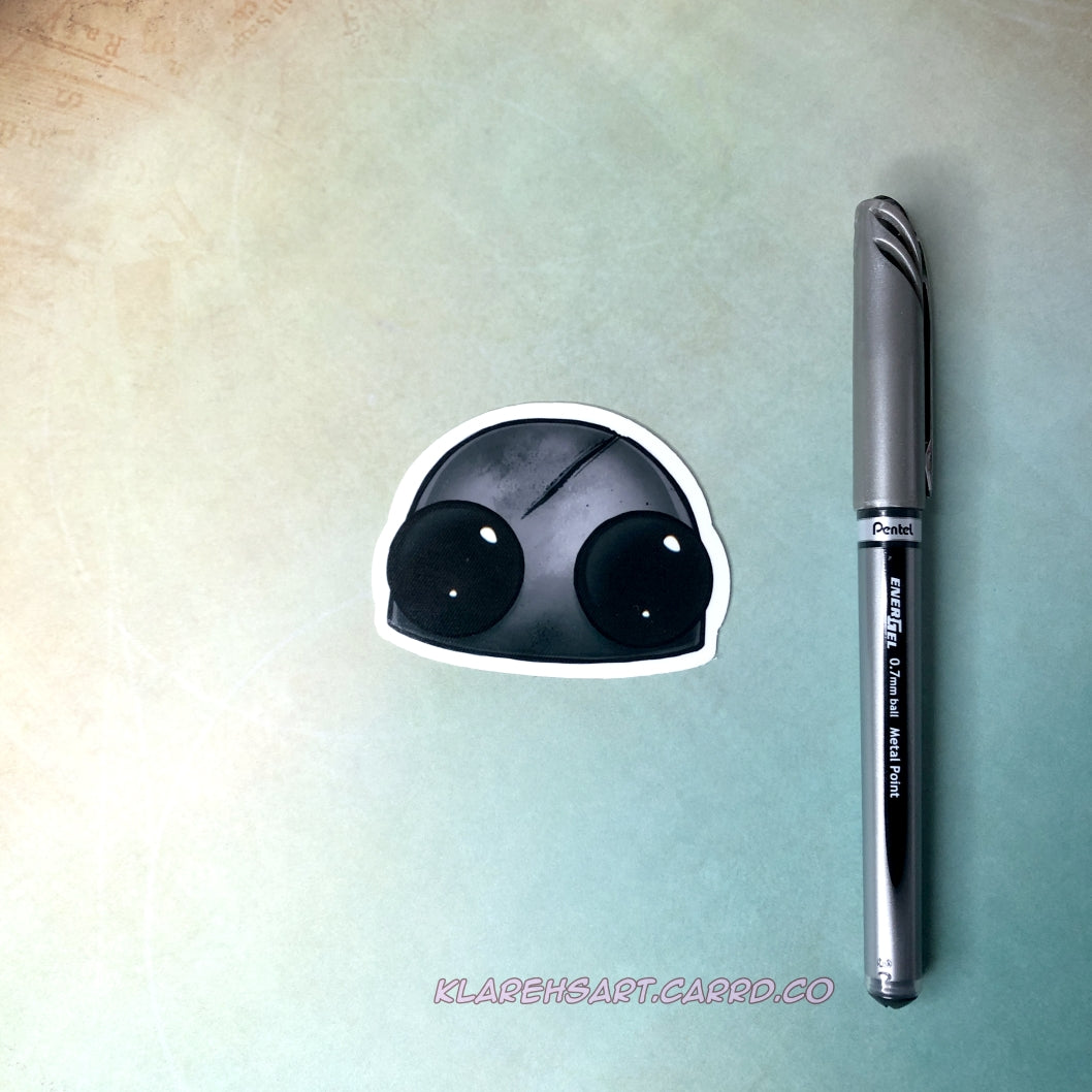 A vinyl sticker of a dead robot head. It's hull is gray and scratched, it's eyes black with no battery. An ink pen is next to it for size comparison. On a light gold and green background.