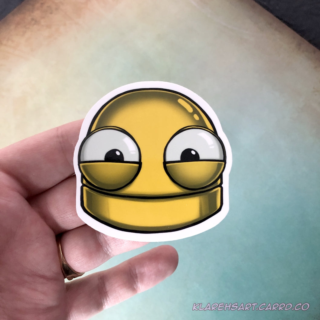 A hand holding a vinyl sticker of a yellow robot head with a happy expression. On a light yellow and green background.