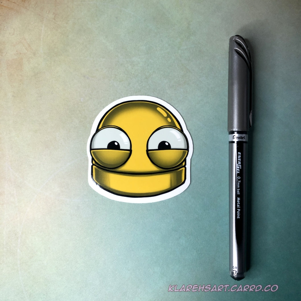 A hand holding a vinyl sticker of a yellow robot head with a happy expression. On a light yellow and green background. An ink pen next to it shows size comparison.