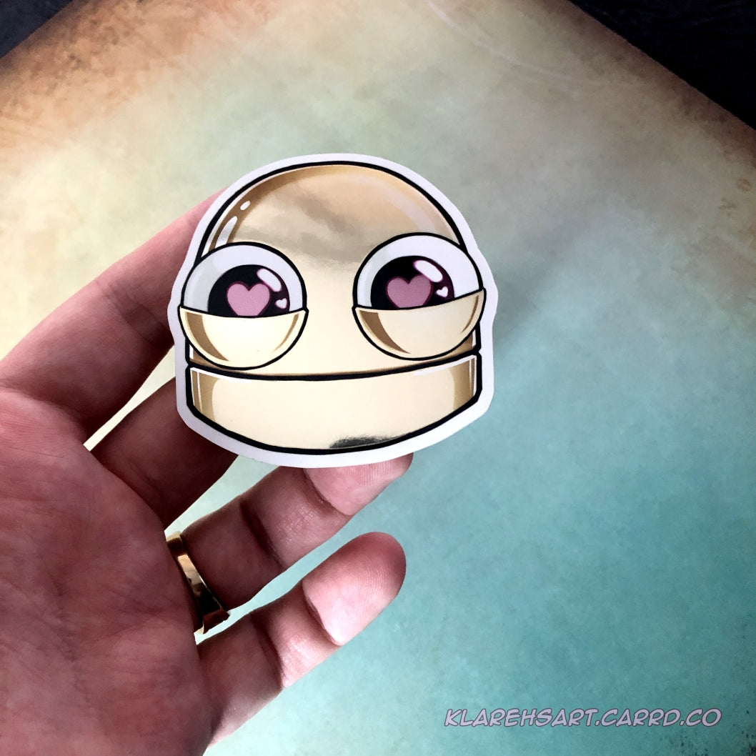 Hand holding a sticker of a gold metallic robot head with heart eyes on a light gold & blue background.
