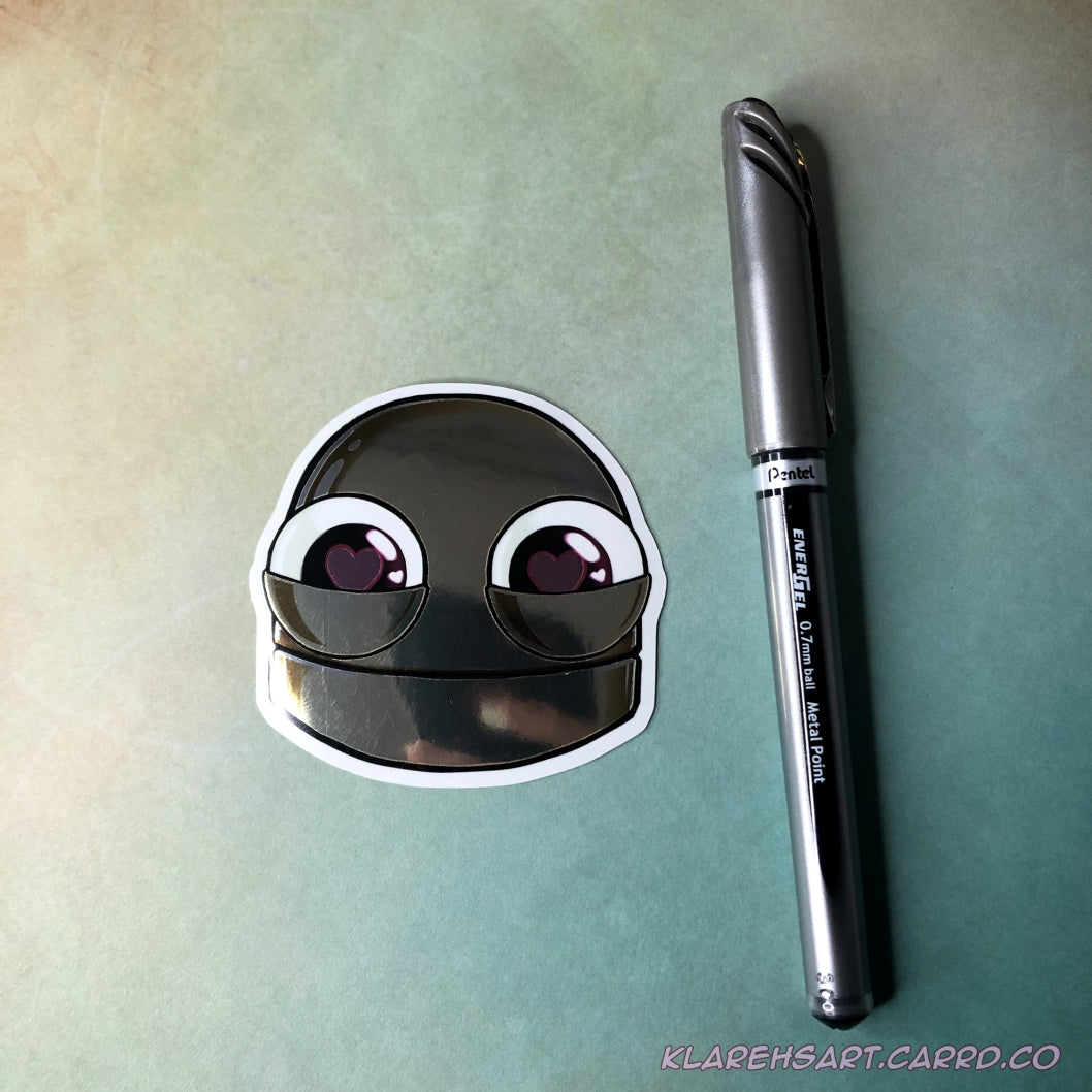 Metallic gold sticker of a  robot head with heart shaped eyes on a light gold and green background, next to an ink pen for size comparison.