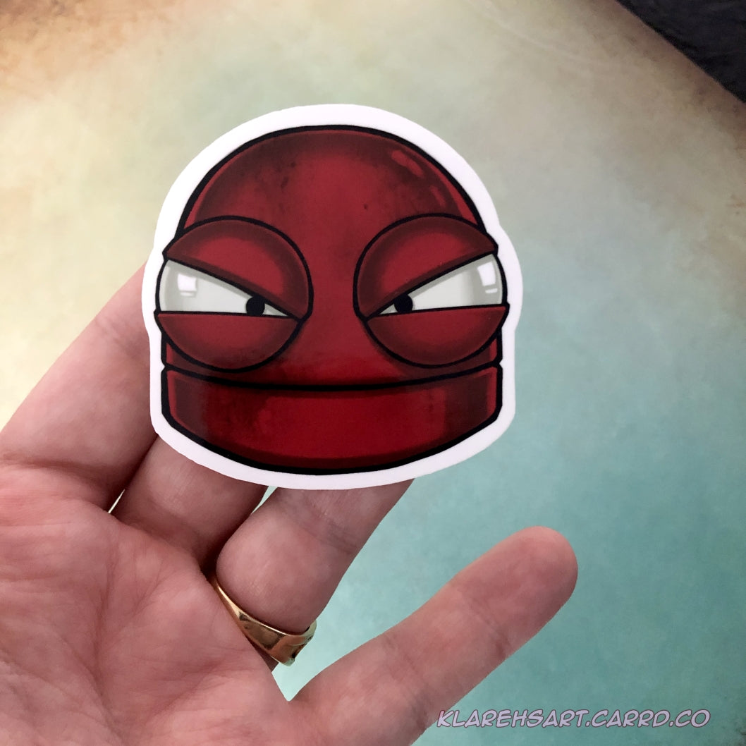 A hand holding a vinyl sticker of a red robot with a mad expression. On a light gold and green background.