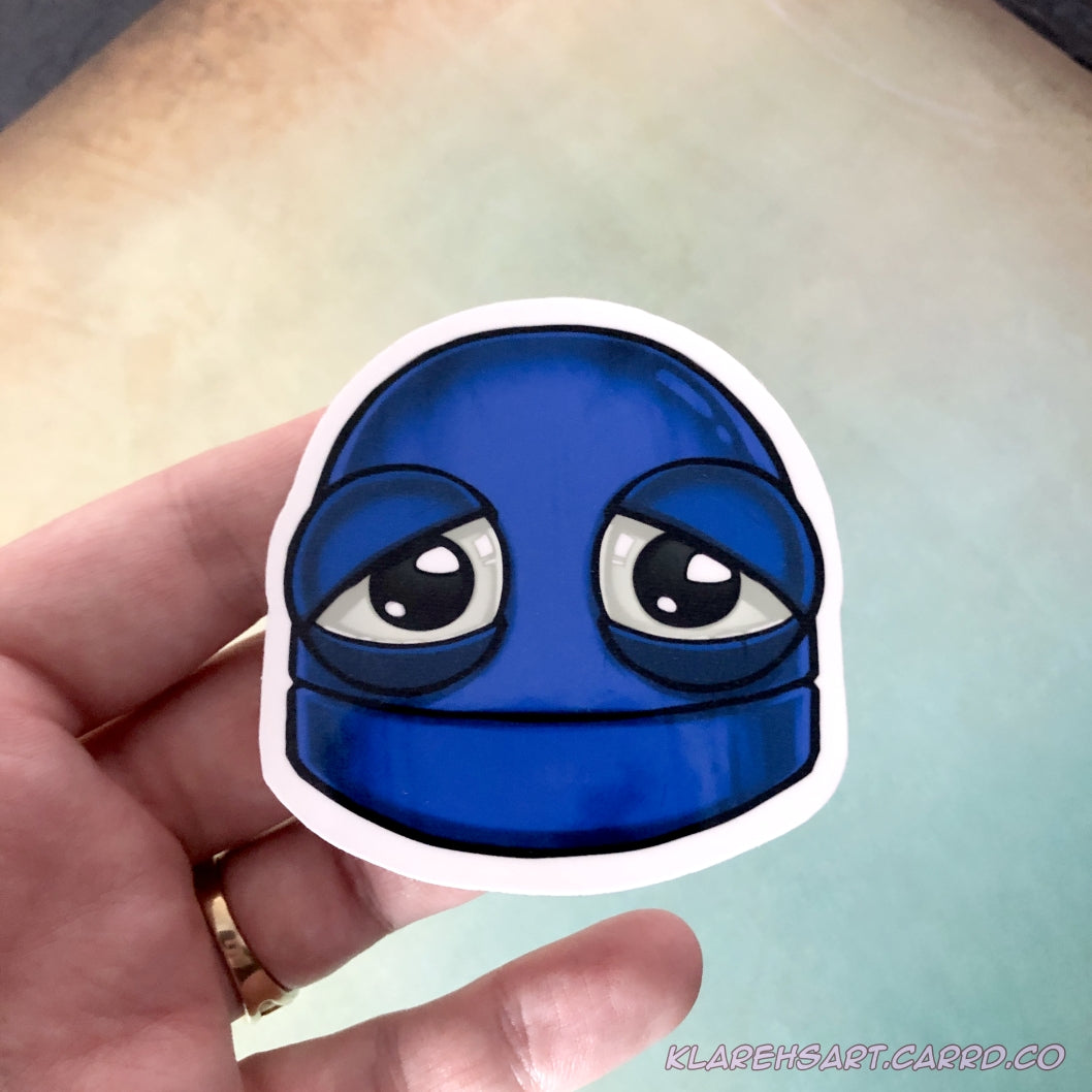 A hand holding a vinyl sticker of a blue robot with a sad expression. It rests on a light gold and green background.