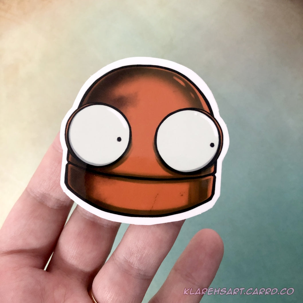 A hand holding an orange vinyl sticker of a robot head. It's eyes wide with fright. It is above a gold & green background.