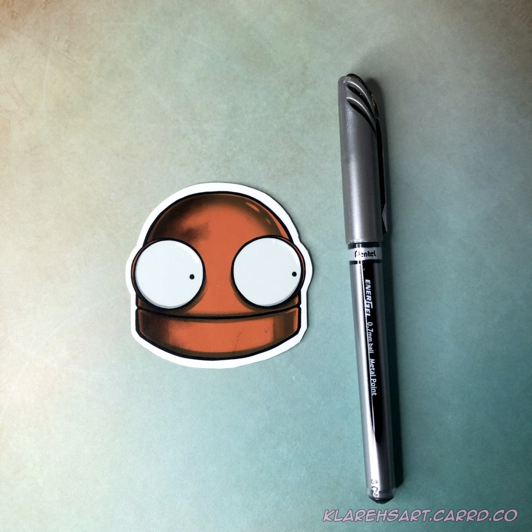 An orange vinyl sticker of a robot head. It's eyes wide with fright. An ink pen next to it is showing size comparison. It is above a gold & green background.