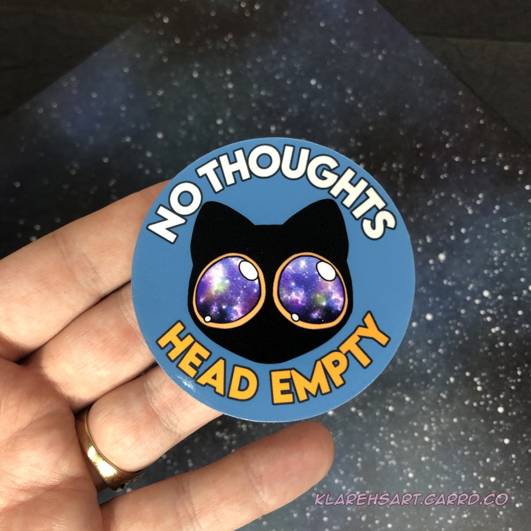 Sticker with a black cat design with galaxy eyes and text 'No Thoughts Head Empty' held by a hand against a starry background.